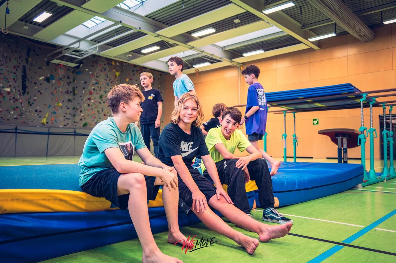Jugendliche beim Ferienprogramm in einer großen Sporthalle — ein Trainer hebt einen Teenager in die Luft, andere Kinder schauen zu